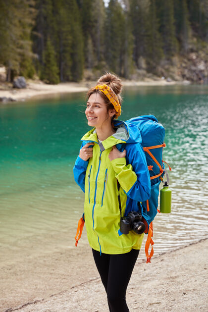 漫步垂直拍摄的旅游女孩走在松石湖 针叶林 微笑着高兴地拿着相机和大背包山娱乐徒步旅行