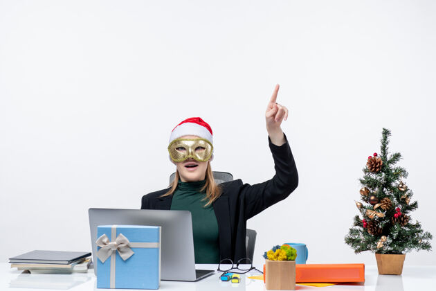 圣诞老人圣诞节的心情与惊讶的年轻女子戴着圣诞老人的帽子 戴着口罩坐在一张桌子上指着白色背景的问题电脑桌子办公室