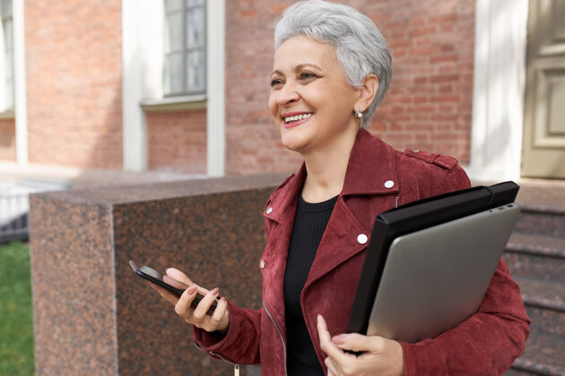 通讯开朗的退休女性在户外摆好姿势 带着便携式电脑和智能手机 笑容灿烂 很高兴见到朋友科技手机时尚