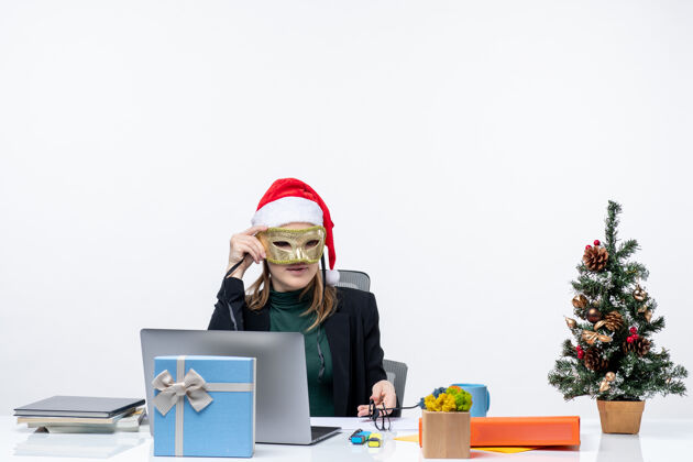 笔记本电脑一位严肃的年轻女士 戴着圣诞老人的帽子 戴着眼镜 坐在一张桌子旁 桌子上有一棵圣诞树和一份白色背景的礼物眼镜圣诞老人认真的