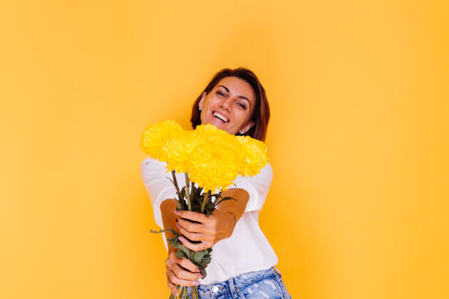 植物摄影棚拍摄的黄色背景快乐的白人妇女短发穿着休闲服白衬衫和牛仔裤手持一束黄色紫苑人气味积极