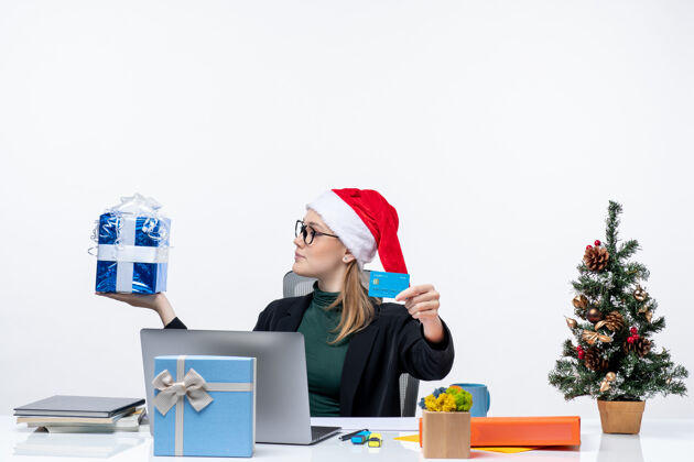 礼物带着圣诞老人帽子戴着眼镜坐在桌子旁拿着圣诞礼物和银行卡的积极的女商人抱着帽子圣诞老人