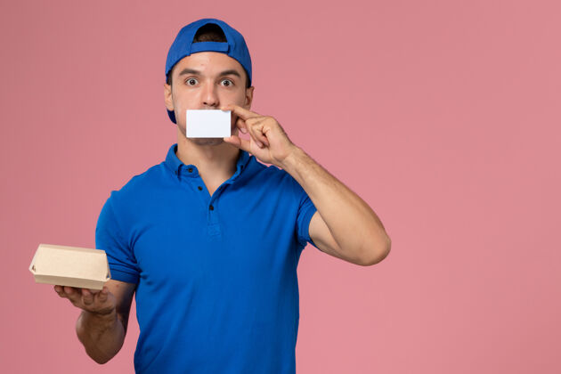 年轻人正面图：身穿蓝色制服披肩的年轻男性信使手拿着小快递食品包和浅粉色墙上的白色卡片食品制服视图