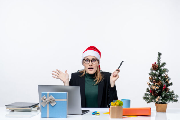 坐着在白色背景的办公室里 一个戴着圣诞老人帽子的金发女人坐在一张桌子旁 桌子上放着一棵圣诞树和一份礼物人帽子金发