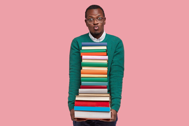 书籍帅气的黑人男子撅着嘴唇 背着课本 对着镜头做鬼脸 穿着绿色的套头衫 对着粉色的墙壁做模特眼镜男性眼镜
