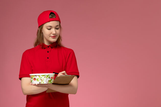 女性正面图：身穿红色制服和披风的年轻女快递员拿着投递碗和记事本在粉色墙上写笔记年轻女性快递人女人