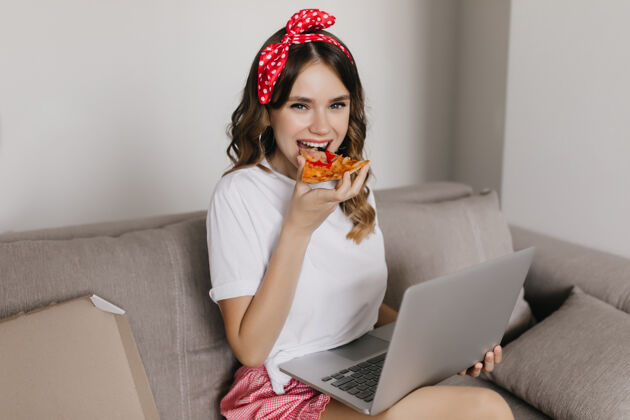 人迷人的卷发女孩愉快地吃比萨饼和使用电脑迷人的女人周末在家里用笔记本电脑和快餐金发互联网沙发