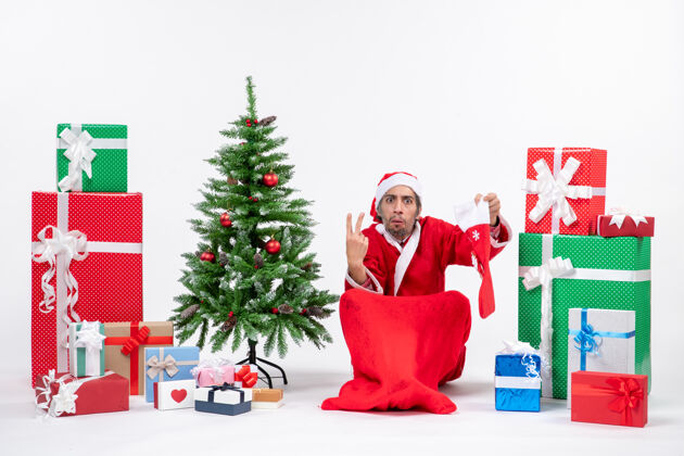 袜子紧张的圣诞老人展示了两个坐在地上拿着圣诞袜的礼物和装饰在白色背景上的新年树圣诞老人装饰圣诞老人