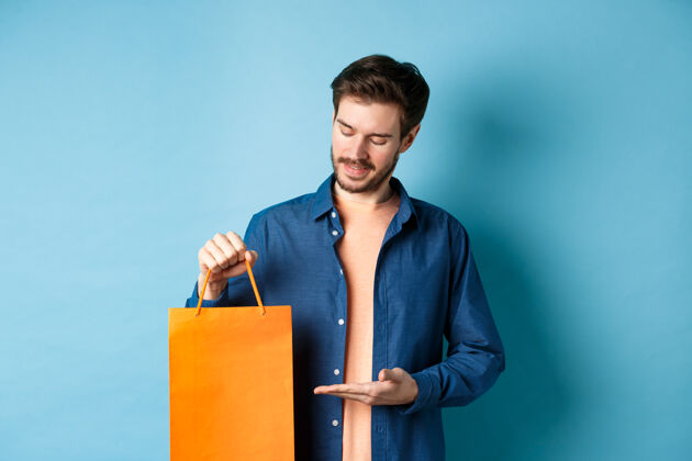 男孩年轻人微笑着展示橙色购物袋 展示礼物 站在蓝色背景上手休闲脸