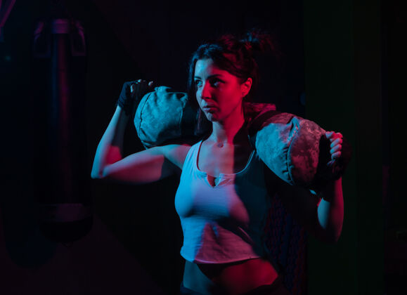 包年轻的运动女性拿着沉重的健身袋在霓虹灯梯度蓝红光下锻炼 在黑暗的墙壁上功能训练概念减肥运动员健身房