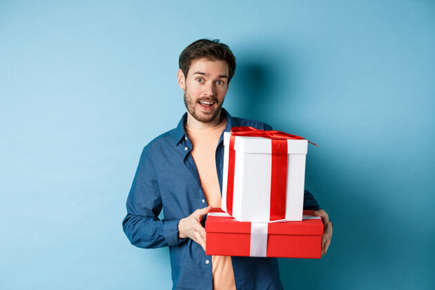 拥抱帅哥留着胡子 拿着情人节礼物 拿着礼物站在盒子里 看着相机 蓝色背景男朋友情人情感