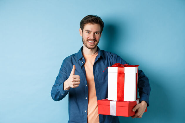 感情微笑的男人拿着浪漫的礼物 竖起大拇指 庆祝情人节 给爱人买礼物 站在蓝色的背景上丈夫温柔情感