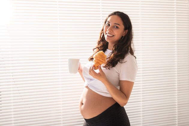 怀孕早上吃早餐时 美丽的孕妇手里拿着牛角包和一杯咖啡女性化责任产妇