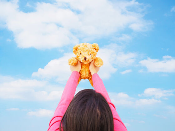 毛绒后视图亚洲女孩与长发举行棕色泰迪熊玩具在蓝天背景童年和快乐的孩子们长女性动物