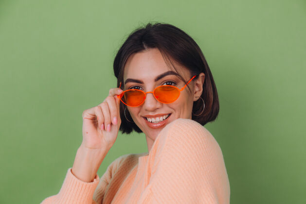 兴奋年轻时尚的女士穿着休闲的桃色毛衣 戴着橙色的眼镜 隔着绿橄榄色的墙壁 开心的 积极的笑着 围着复制空间转女人情感衣服