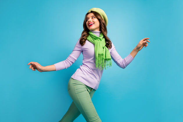 乐趣低视角下迷人美女的侧面照片旅行者走在街上兴奋的心情戴绿色贝雷帽紫色套头衫围巾裤子孤立的蓝色墙壁个人资料裤子围巾