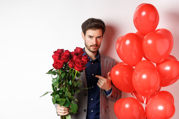 男人穿着西装的帅哥在浪漫的情人节约会 指着一束玫瑰花 站在白色背景上的心气球旁玫瑰感情伴侣