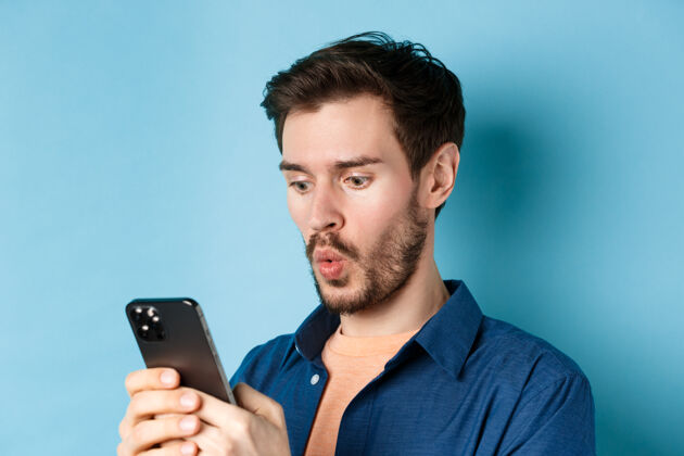 手一个年轻人看着智能手机说哇哦 查看网上的报价 站在蓝色的背景上表情小玩意发型