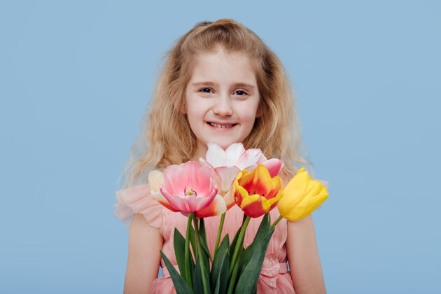 快乐微笑着的小女孩 穿着粉红色的裙子 手里拿着春天的花朵 孤零零地站在蓝色的墙上看微笑童年