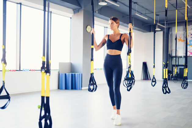 肌肉美丽的年轻女子摆姿势与悬挂教练吊带或吊带在健身房教练运动员体重