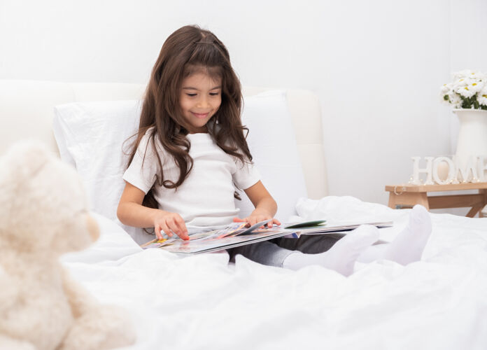 教育快乐的小黑发女孩坐在家里的床上看书家庭房子玩具
