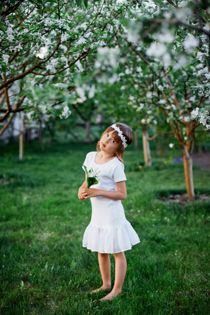 卷发可爱的5-6岁小女孩手持鲜花站在百花齐放的春天花园里 穿着白色的连衣裙 在户外戴着花环 春天来了年轻生活方式休闲