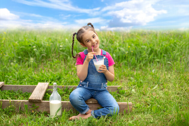 玻璃一个快乐的女孩扎着辫子 穿着牛仔工作服和粉色t恤 手里拿着一杯牛奶和一根粉色的吸管 坐在一个木梯子上 在草地上的田野里蓝天白云复制空间饮料颜色乡村