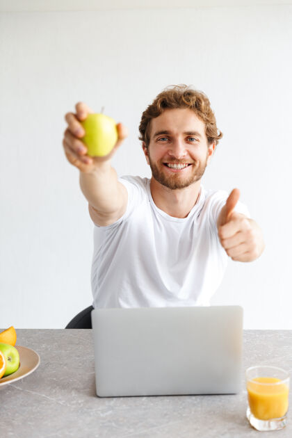 舒适快乐的小胡子男人在家里的桌子旁用笔记本电脑拿着水果摆出竖起大拇指的手势水果早餐吸引