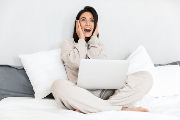 笔记本电脑一张30多岁的兴奋女人在舒适的公寓里用笔记本电脑 穿着白色亚麻布坐在床上的照片可爱成熟白种人