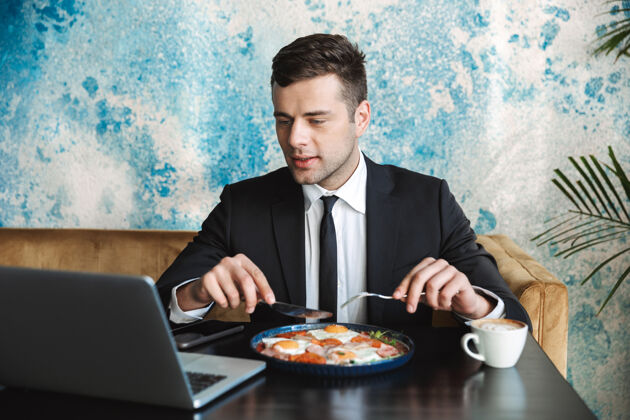 吸引力一个英俊的年轻商人坐在咖啡馆里用笔记本电脑吃早餐或晚餐的画面西装咖啡馆企业家