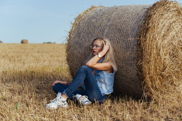 田野一位年轻漂亮的女村民穿着牛仔裤在一捆干草旁摆姿势稻草美丽干草