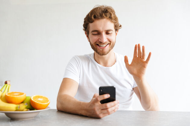 软件照片中 一个快乐的留着胡子的年轻人在家里的桌子旁用手机挥手交谈笔记本电脑通话休闲