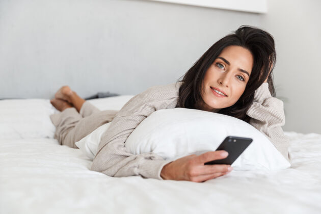 可爱一张30多岁的熟女在明亮的房间里用手机躺在白色亚麻布的床上的照片舒适互动中年