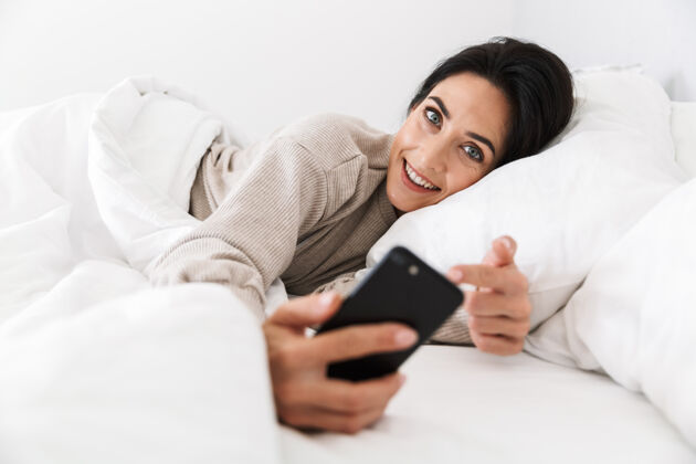 沙发30多岁的快乐女人在家里穿着白色亚麻布躺在床上使用智能手机的照片互动可爱公寓