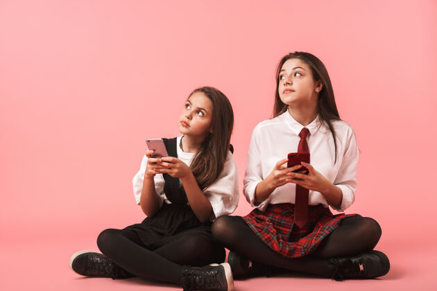 积极身着校服的快乐女孩们一边用手机 一边坐在隔着红墙的地板上电话姿势请