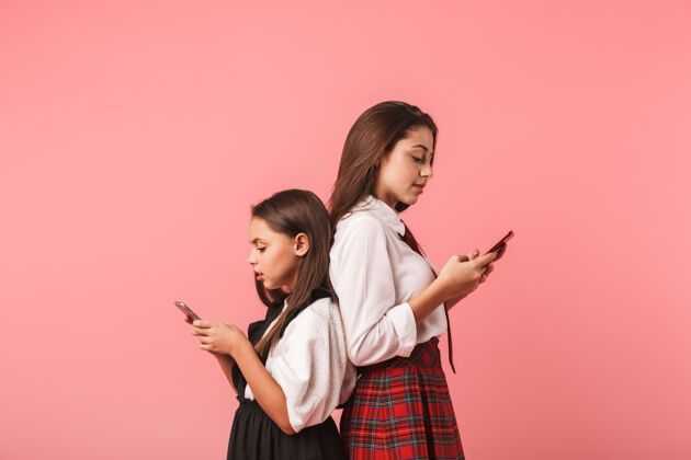 课程身着校服的黑发女孩使用智能手机的肖像 同时孤立地站在红墙上制服姿势青少年