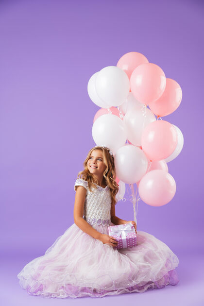 孩子穿着公主裙的漂亮小女孩孤零零地坐在紫罗兰色的墙上 手里拿着一堆气球和一个礼盒童年爱小