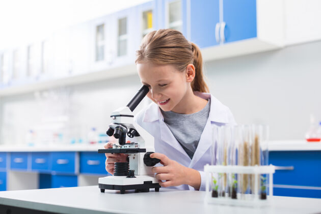 专业知识怎样令人惊奇愉快高兴的女孩微笑着调显微镜 穿着实验服解决方案尼斯实验室外套
