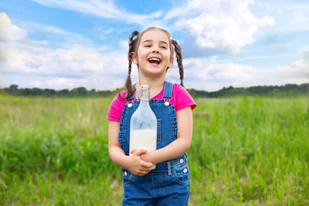 田野在草地上的田野里 一个梳着辫子 穿着牛仔工作服和粉色t恤 手里拿着一瓶牛奶的欢笑女孩蓝天白云吃生活方式一天
