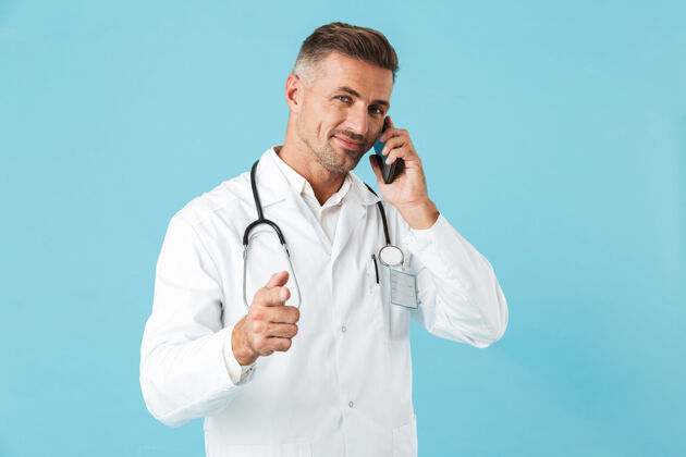 从业者积极的医生穿着白大褂和听诊器讲手机的照片 孤立地站在蓝色的墙上讲话成人微笑