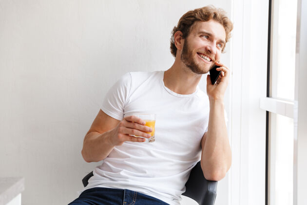 男士微笑的年轻人在讲手机 看着家里的窗户 喝着橙汁手机快乐交谈