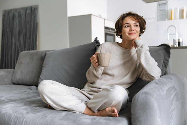 年轻快乐的年轻女子坐在家里的沙发上 手里拿着杯子茶休息白种人