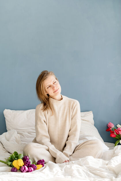 庆祝快乐的女人穿着睡衣坐在床上捧着郁金香花束 背景是蓝色的墙壁床休闲房间