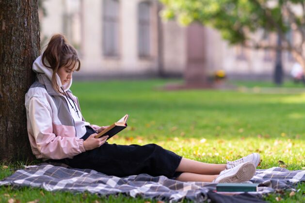 地毯在公园或花园的树荫下看书或学习的年轻女子 近距离的侧视沉浸爱好活动