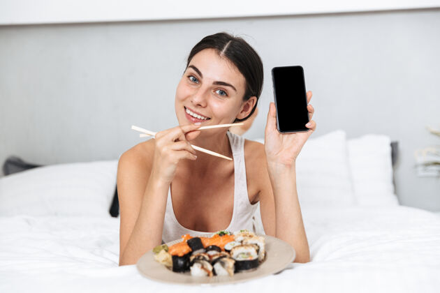 兴趣美丽的年轻女子在家里的床上放松 拿着手机 吃着盘子里的寿司放松卧室交流