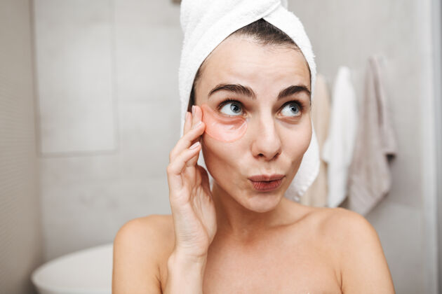 触摸特写镜头：一个头上戴着毛巾的漂亮年轻女子站在浴室里 敷眼罩头部保湿皮肤护理