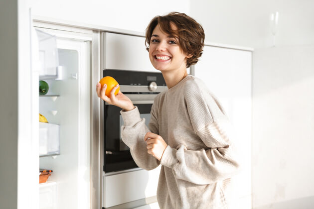 新鲜微笑的年轻女子站在冰箱前 手里拿着橘子概念膳食小吃