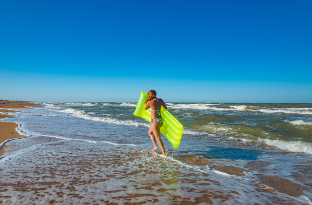 美丽穿着泳装的漂亮苗条的年轻女子拿着气垫在沙滩上奔跑女性泡沫热带
