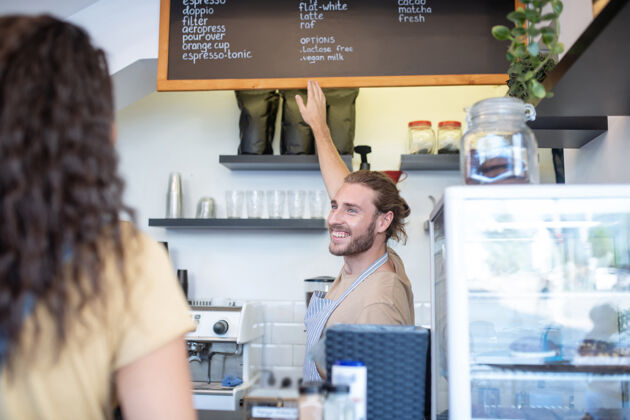 小企业咖啡 咖啡选择快乐围裙上留着胡子的年轻人指着菜单 墙上挂着各种各样的咖啡 后面是女人自营职业销售选择