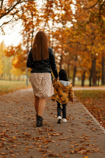 人行道公园里一个穿着时尚服饰的年轻女子和一个穿着时尚服饰的孩子的后视图妈妈美丽秋天